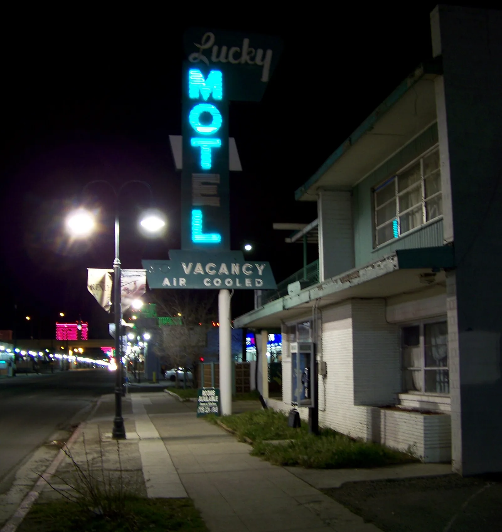 A tall neon Lucky Motel sign glowing blue with a Vacancy panel, beside a two-story motel on a city street at night