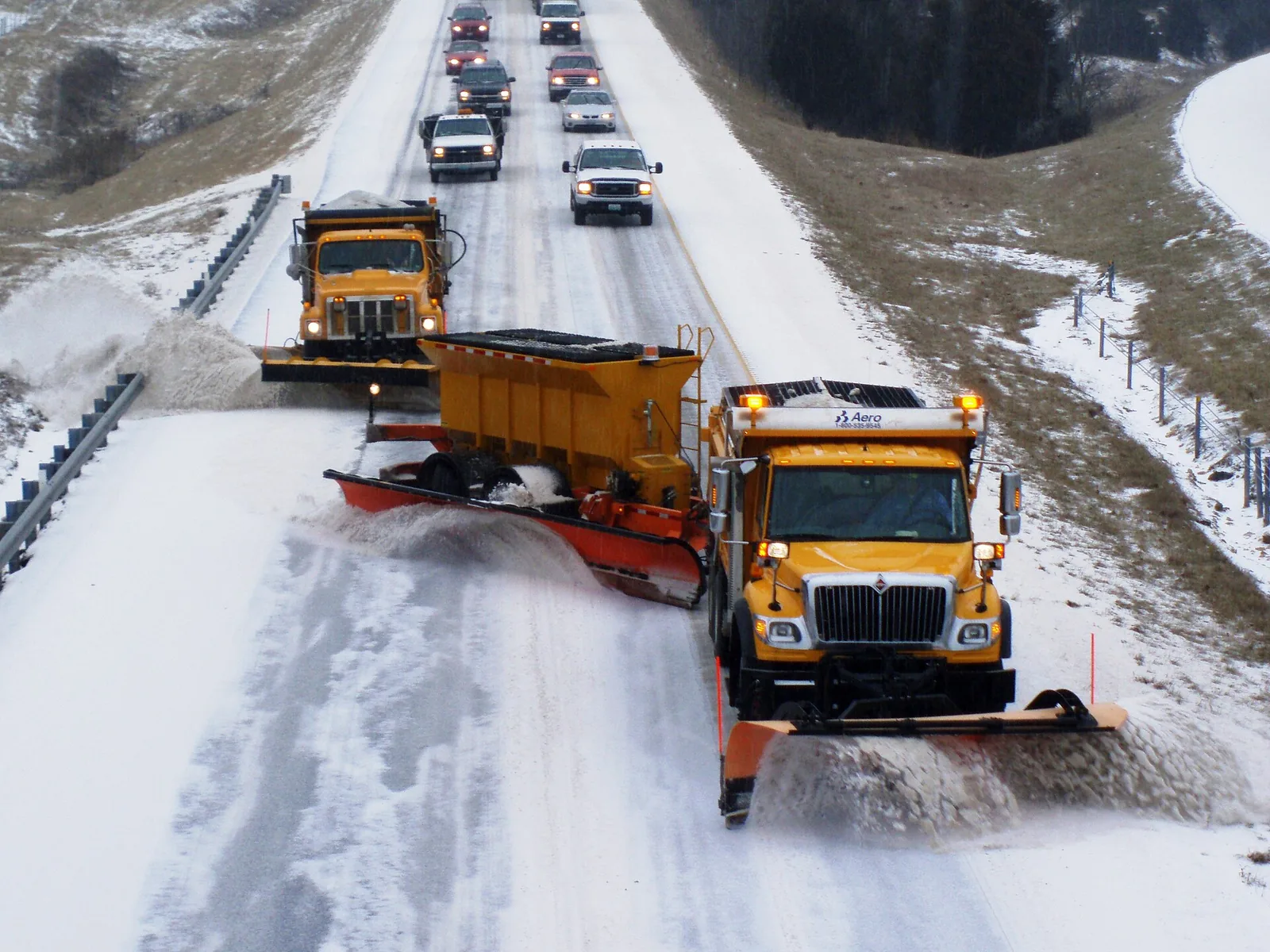 Two orange-and-yellow snowplows, one towing a second plow blade, clearing a snowy rural interstate in Missouri with cars following behind
