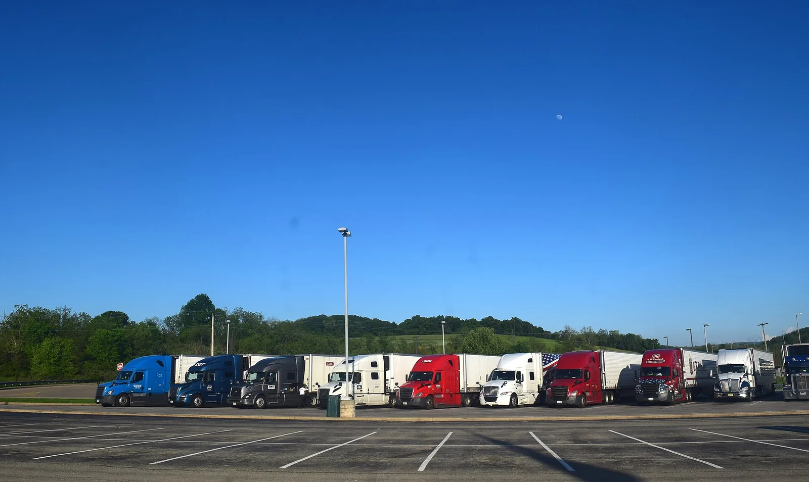 A row of tractor-trailers parked side by side in a large paved lot beside a highway under a clear sky